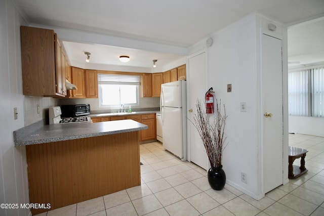 kitchen featuring a peninsula, light tile patterned floors, freestanding refrigerator, under cabinet range hood, and a sink