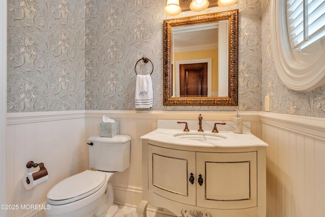half bath featuring toilet, vanity, wallpapered walls, and a wainscoted wall