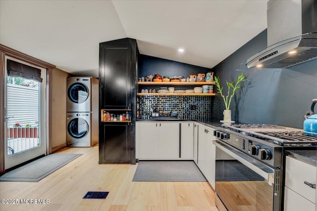 kitchen with stacked washer / drying machine, open shelves, wall chimney range hood, gas stove, and dark countertops