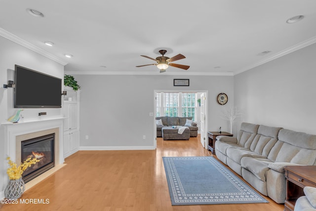 living area with a glass covered fireplace, baseboards, light wood-type flooring, ornamental molding, and a ceiling fan