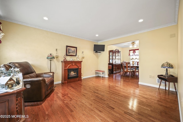 living room with a warm lit fireplace, wood finished floors, a ceiling fan, crown molding, and baseboards