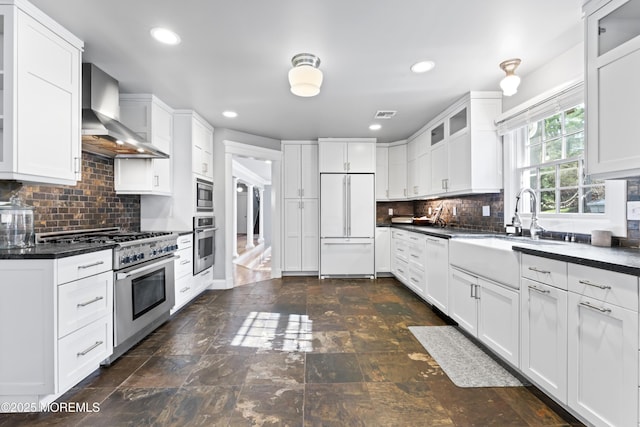 kitchen featuring glass insert cabinets, built in appliances, a sink, wall chimney range hood, and dark countertops