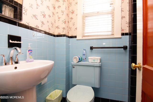 half bath featuring toilet, tile walls, wallpapered walls, a wainscoted wall, and a sink