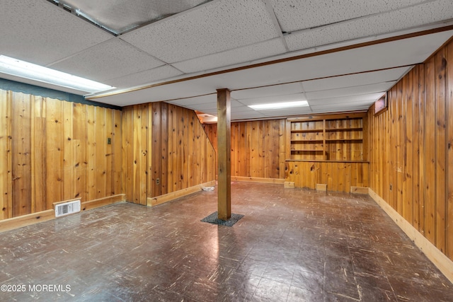 finished below grade area featuring visible vents, baseboards, a drop ceiling, wooden walls, and tile patterned floors