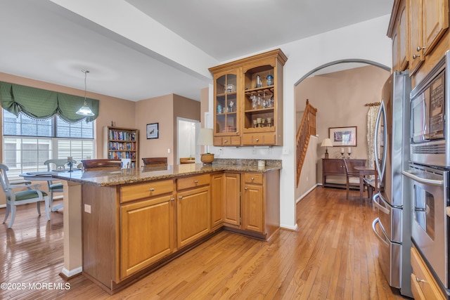 kitchen with glass insert cabinets, appliances with stainless steel finishes, a peninsula, light wood finished floors, and light stone countertops