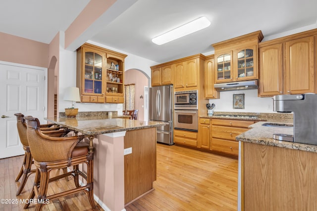 kitchen featuring arched walkways, a peninsula, stone counters, appliances with stainless steel finishes, and under cabinet range hood