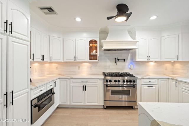 kitchen with light stone countertops, custom exhaust hood, visible vents, white cabinets, and appliances with stainless steel finishes
