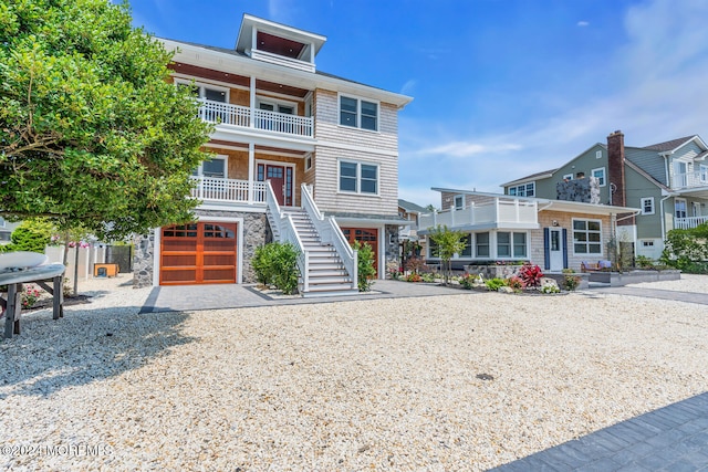 coastal home featuring stone siding, an attached garage, stairs, a balcony, and gravel driveway