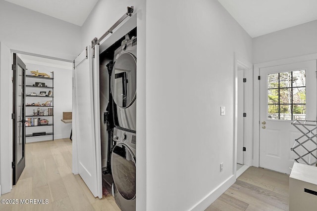 laundry area featuring laundry area, a barn door, light wood finished floors, stacked washing maching and dryer, and baseboards