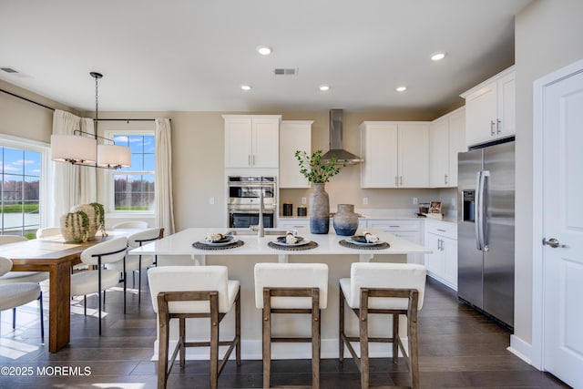 kitchen featuring appliances with stainless steel finishes, dark wood-style flooring, wall chimney range hood, light countertops, and an island with sink