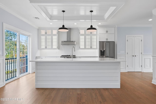 kitchen featuring wall chimney range hood, wood finished floors, a kitchen island with sink, stainless steel fridge with ice dispenser, and a tray ceiling
