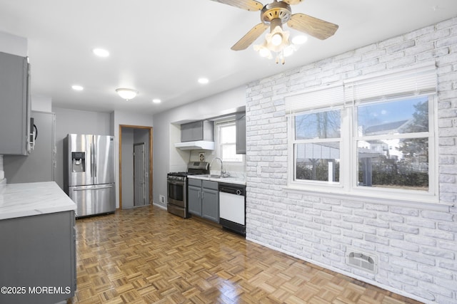 kitchen featuring wall chimney range hood, a ceiling fan, brick wall, stainless steel appliances, and gray cabinetry