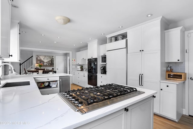kitchen with light stone counters, built in appliances, a sink, white cabinetry, and light wood-type flooring