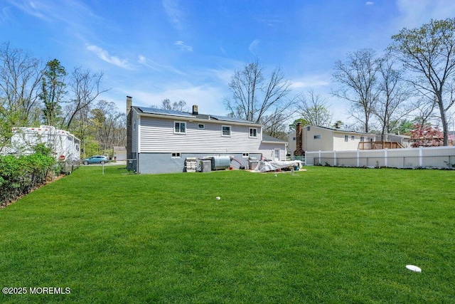 rear view of house featuring a chimney, fence, roof mounted solar panels, and a lawn
