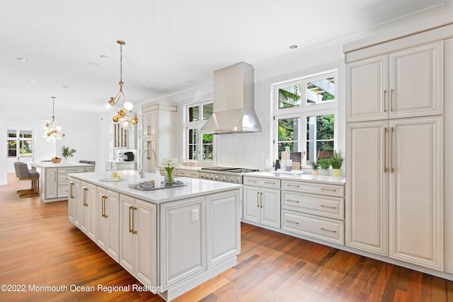 kitchen with wall chimney range hood, light countertops, light wood finished floors, an island with sink, and a notable chandelier