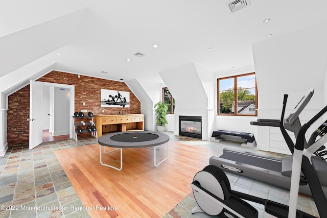 workout room featuring a glass covered fireplace, visible vents, brick wall, and vaulted ceiling
