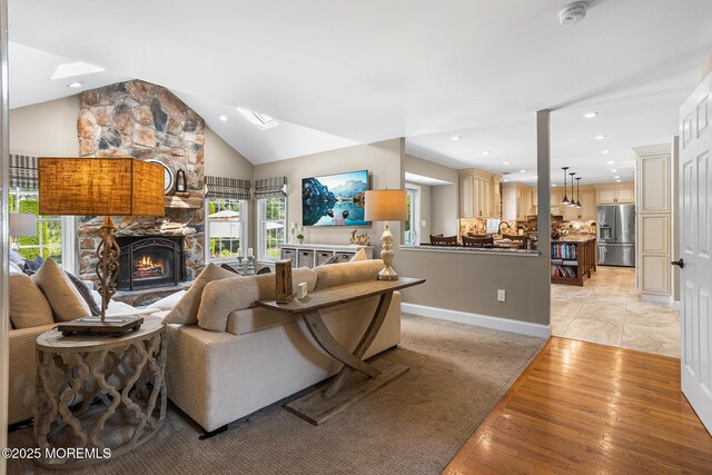 living room with vaulted ceiling with skylight, a stone fireplace, light wood-style flooring, recessed lighting, and baseboards