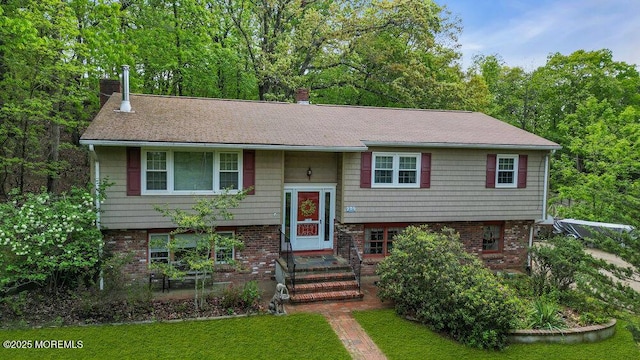 raised ranch featuring a chimney, a shingled roof, brick siding, and a front lawn