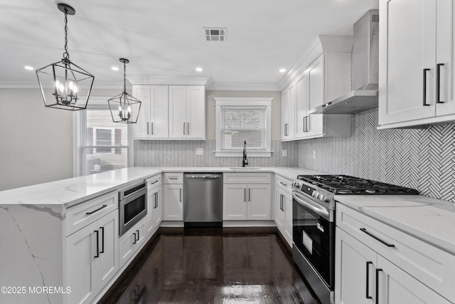 kitchen featuring a peninsula, wall chimney range hood, a sink, a notable chandelier, and stainless steel appliances