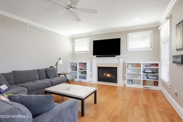 living room with a ceiling fan, visible vents, a fireplace with flush hearth, light wood-style floors, and ornamental molding