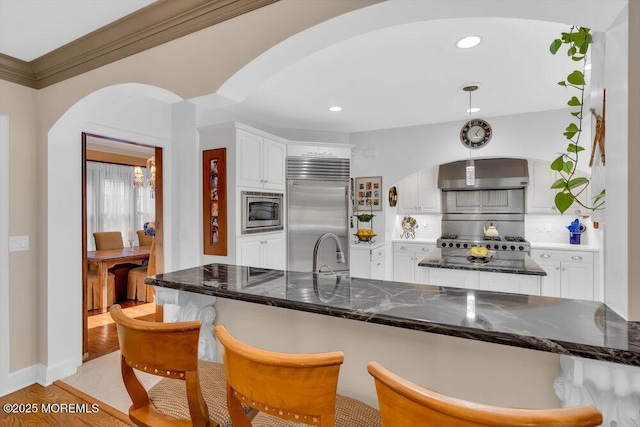 kitchen with arched walkways, decorative backsplash, built in appliances, white cabinets, and a kitchen bar