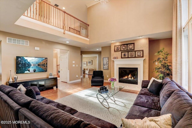 living room with a glass covered fireplace, baseboards, wood finished floors, a towering ceiling, and visible vents