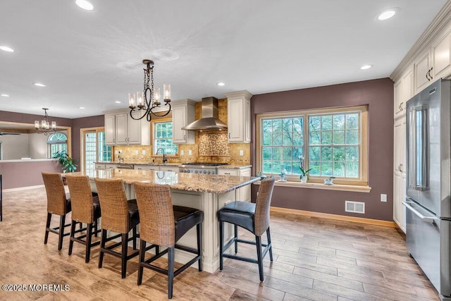 kitchen with wall chimney range hood, a sink, baseboards, a chandelier, and stainless steel fridge
