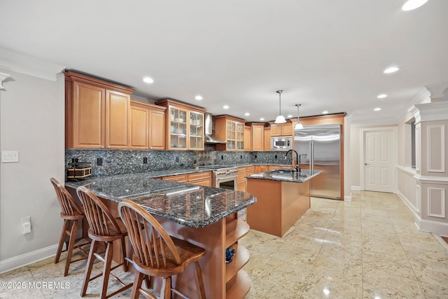kitchen featuring built in appliances, wall chimney range hood, baseboards, an island with sink, and backsplash