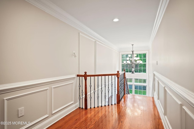 hall with a decorative wall, a chandelier, ornamental molding, wood finished floors, and an upstairs landing