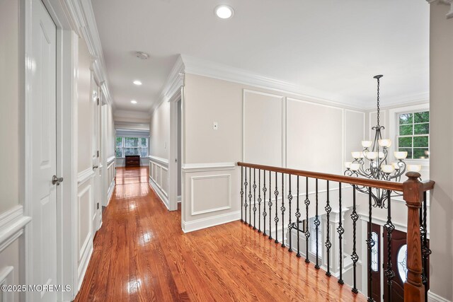 corridor featuring a decorative wall, a chandelier, light wood-style flooring, crown molding, and recessed lighting