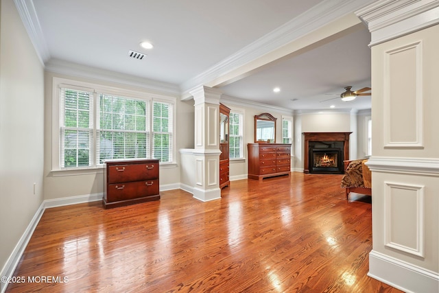 living area featuring ornate columns, ceiling fan, a warm lit fireplace, wood finished floors, and crown molding