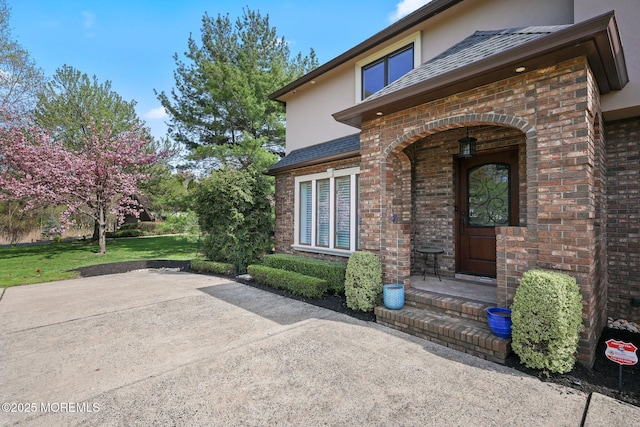 view of exterior entry with brick siding, a lawn, stucco siding, and a patio area