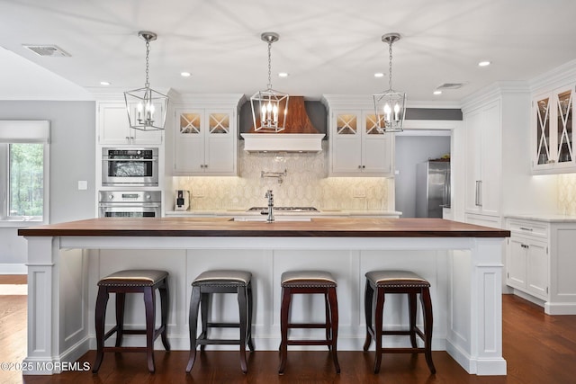 kitchen featuring visible vents, butcher block counters, glass insert cabinets, custom range hood, and stainless steel appliances