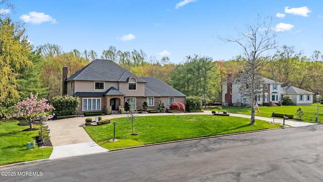 french provincial home with stucco siding, a front yard, a view of trees, a chimney, and concrete driveway