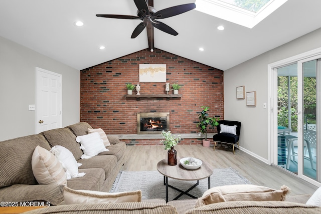 living area featuring a ceiling fan, a fireplace, baseboards, lofted ceiling with skylight, and light wood-style floors