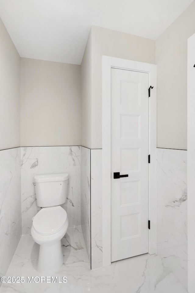 half bathroom with toilet, tile walls, marble finish floor, and a wainscoted wall