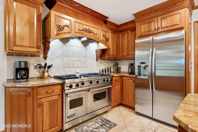 kitchen with decorative backsplash, light tile patterned flooring, brown cabinets, custom range hood, and appliances with stainless steel finishes