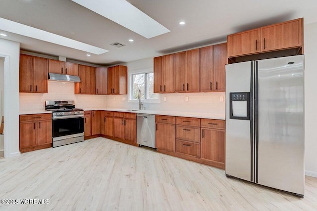 kitchen with appliances with stainless steel finishes, light countertops, brown cabinetry, light wood-type flooring, and under cabinet range hood