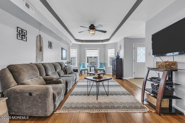 living area with visible vents, wood finished floors, baseboards, a tray ceiling, and a ceiling fan