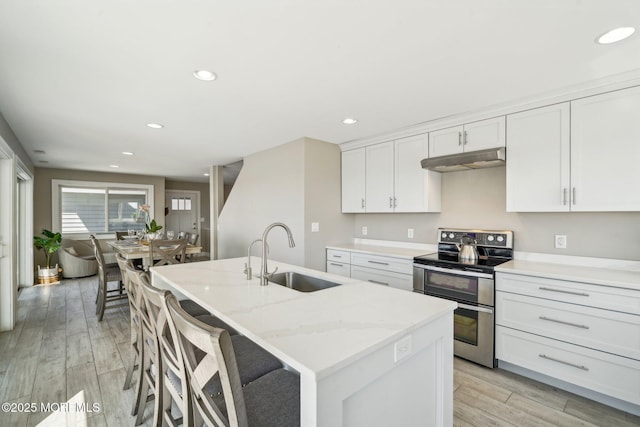 kitchen with light wood-style floors, an island with sink, under cabinet range hood, range with two ovens, and a sink