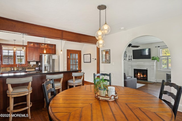 dining space featuring dark wood-style flooring, a brick fireplace, arched walkways, and a ceiling fan