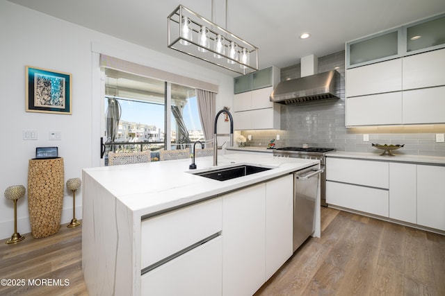 kitchen with wall chimney range hood, decorative backsplash, stainless steel dishwasher, wood finished floors, and a sink