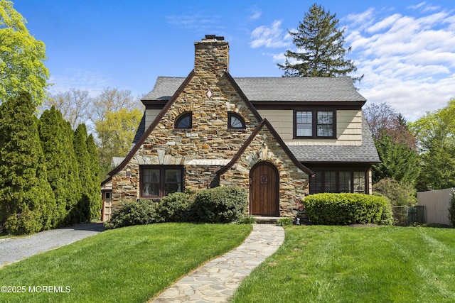 tudor house featuring a front lawn, fence, stone siding, a chimney, and a shingled roof