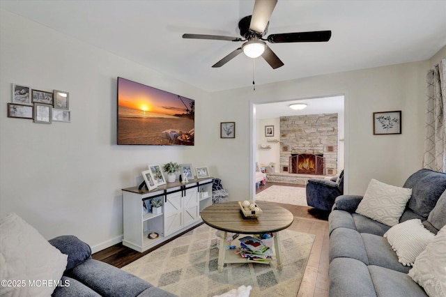 living room featuring baseboards, ceiling fan, a stone fireplace, and wood finished floors