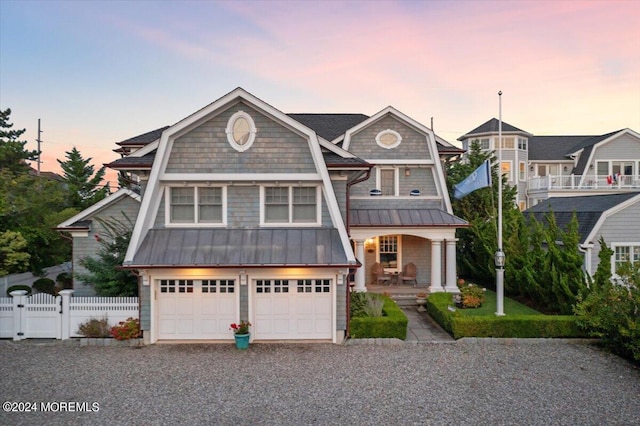 shingle-style home with covered porch, a standing seam roof, fence, gravel driveway, and an attached garage