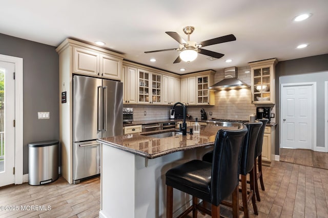 kitchen featuring wall chimney range hood, a ceiling fan, cream cabinets, high quality fridge, and a sink