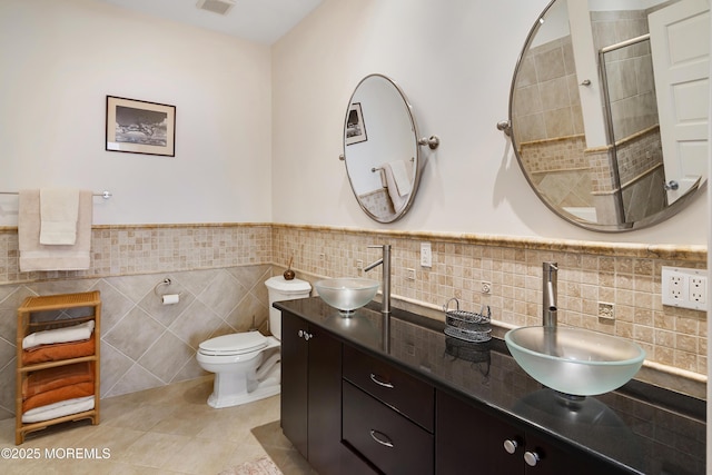 bathroom with tile walls, toilet, a tile shower, double vanity, and wainscoting