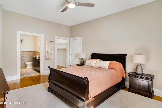 bedroom with wood finished floors, ensuite bath, baseboards, a walk in closet, and ceiling fan