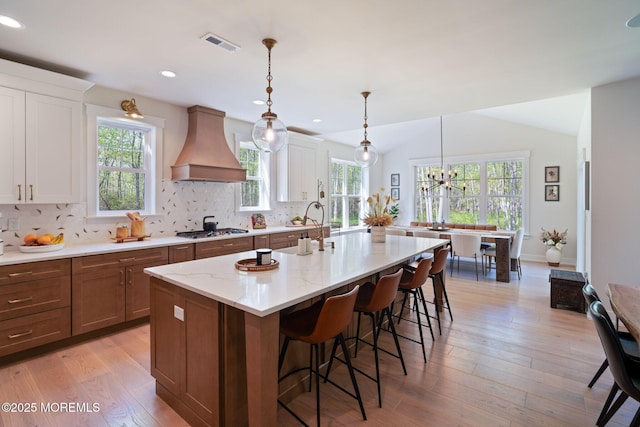 kitchen with custom exhaust hood, stainless steel gas cooktop, a kitchen island with sink, a kitchen breakfast bar, and tasteful backsplash