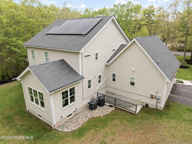 back of house featuring cooling unit, solar panels, roof with shingles, and a lawn
